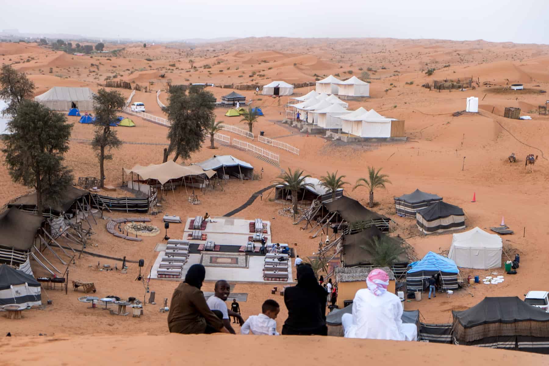 A small gathering of five people sit at the top of a golden sand hill overlooking a tented Bedouin Camp in Ras Al Khaimah 