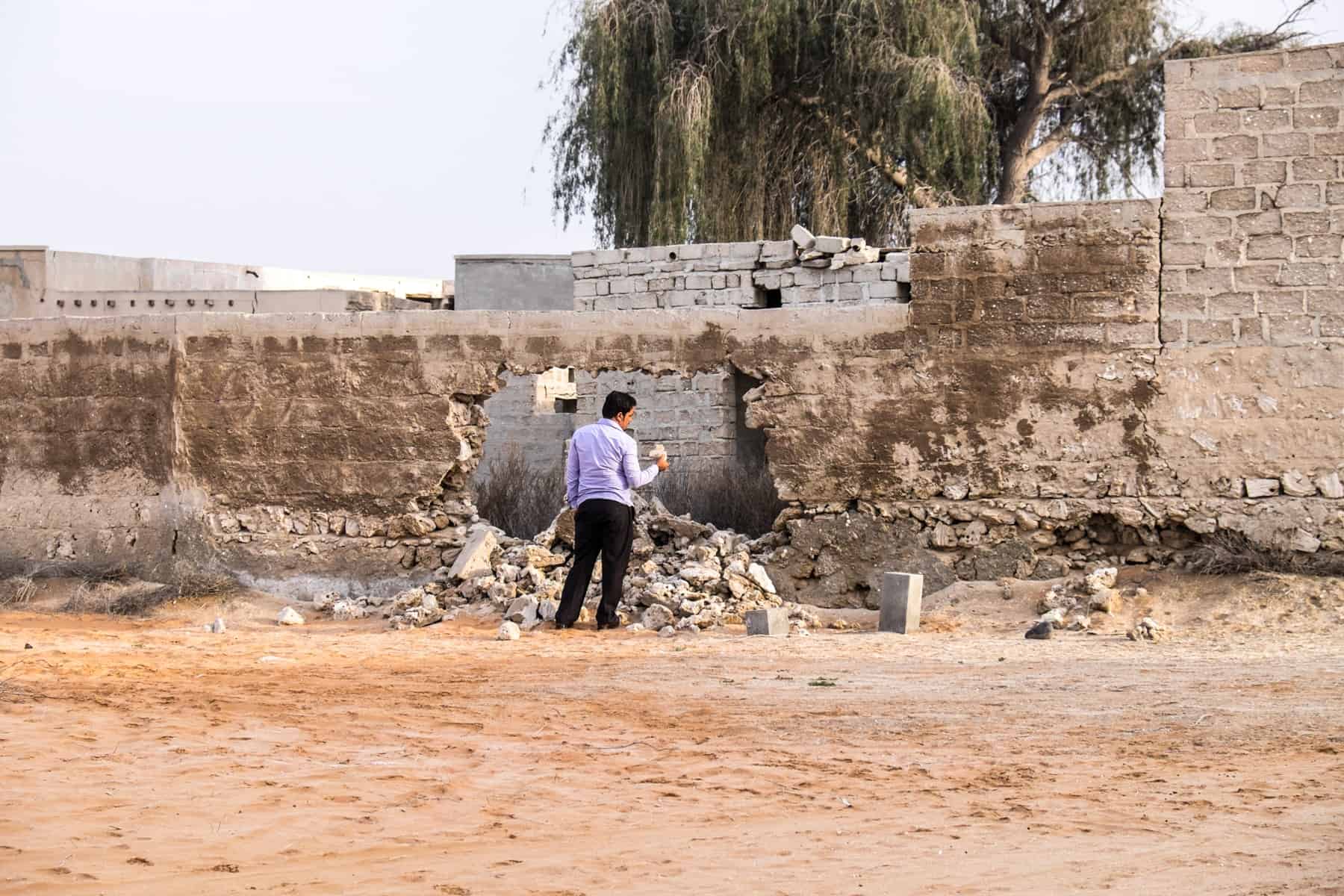 A man in a purple shirt and black trousers looks as a fragment of rock from a crumbling sandy wall in Al Jazirat Al Hamra ghost town in Ras Al Khaimah 