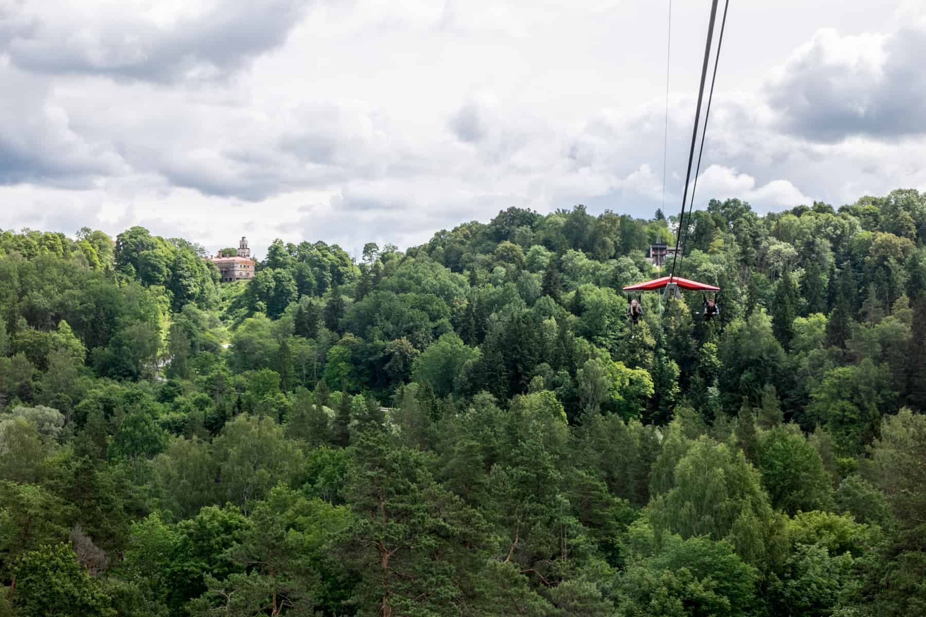 Two people glide through the air on a zipline against a backdrop of dense forest and a castle in Sigulda, Latvia