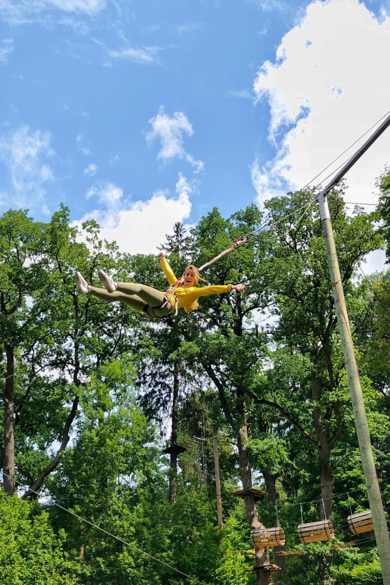 A woman in green jeans and yellow jacket is suspended mid air against a backdrop of trees, happily screaming on a cataput line at an adventure park