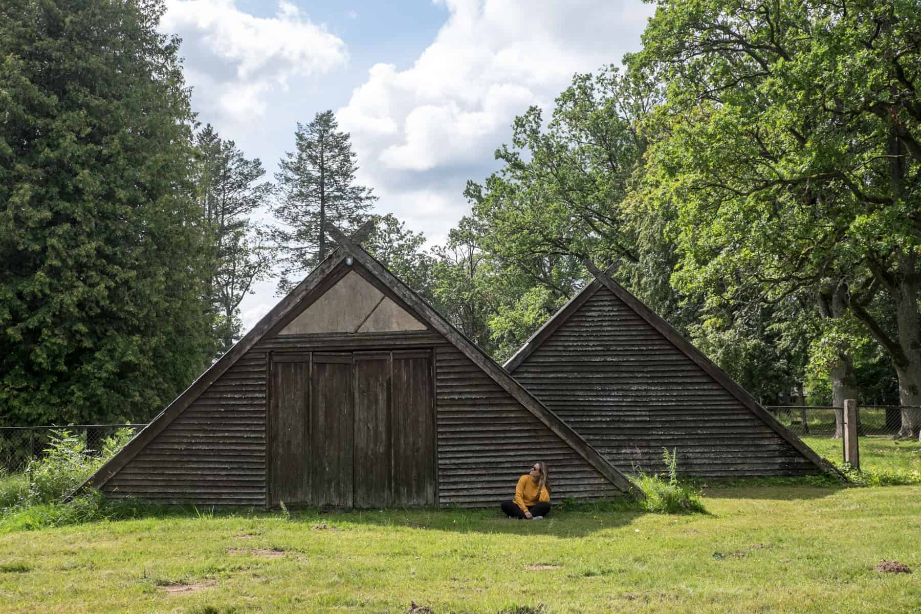 A woman in a yellow jumps sits on the grass in front of a brown, wooden triangular building, in the parkland of Sigulda, Latvia 
