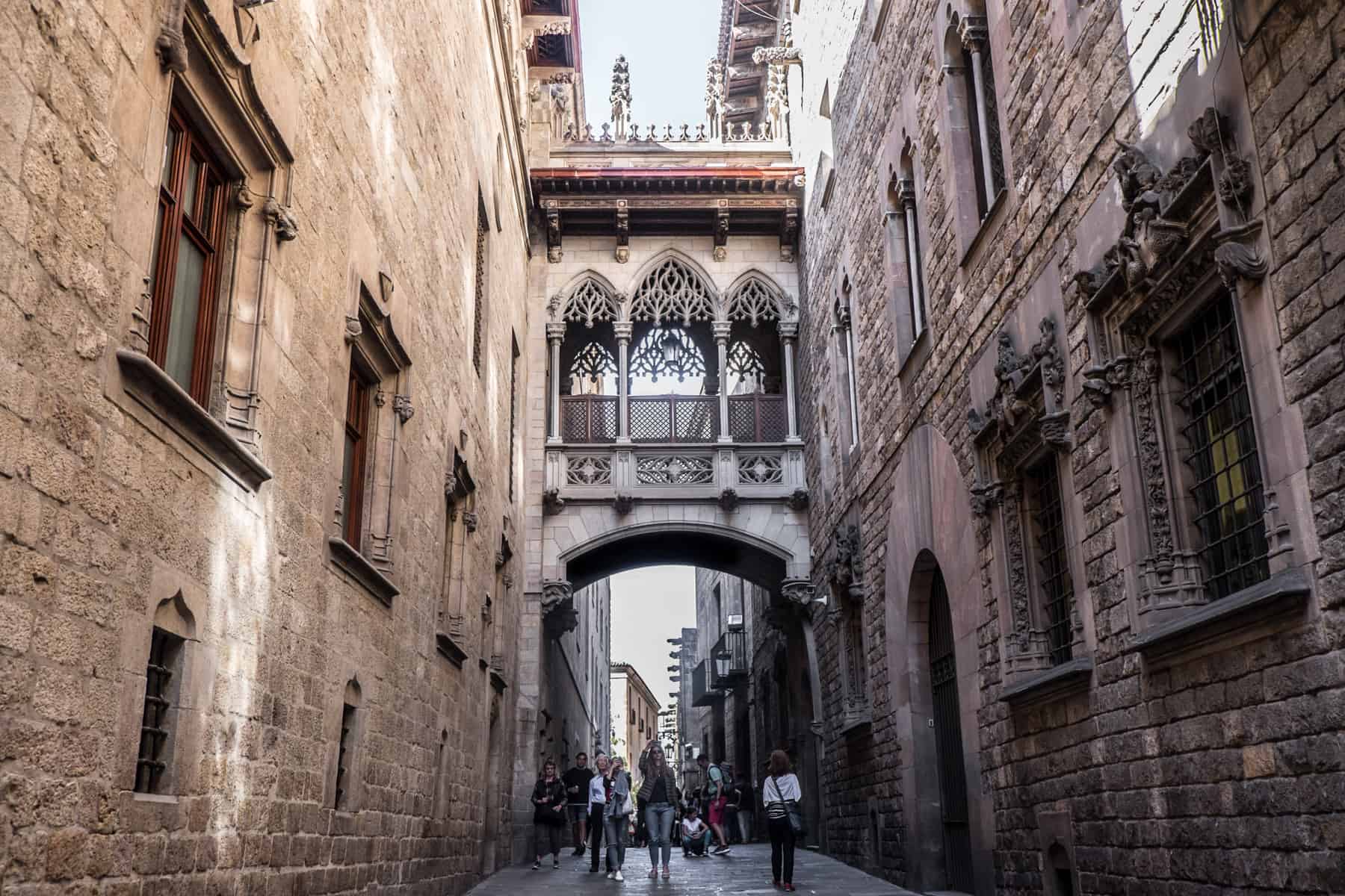 A narrow, delicately carved balcony connects two old golden and brown stone buildings on a long street in Barcelona. People are walking underneath the structure. . 