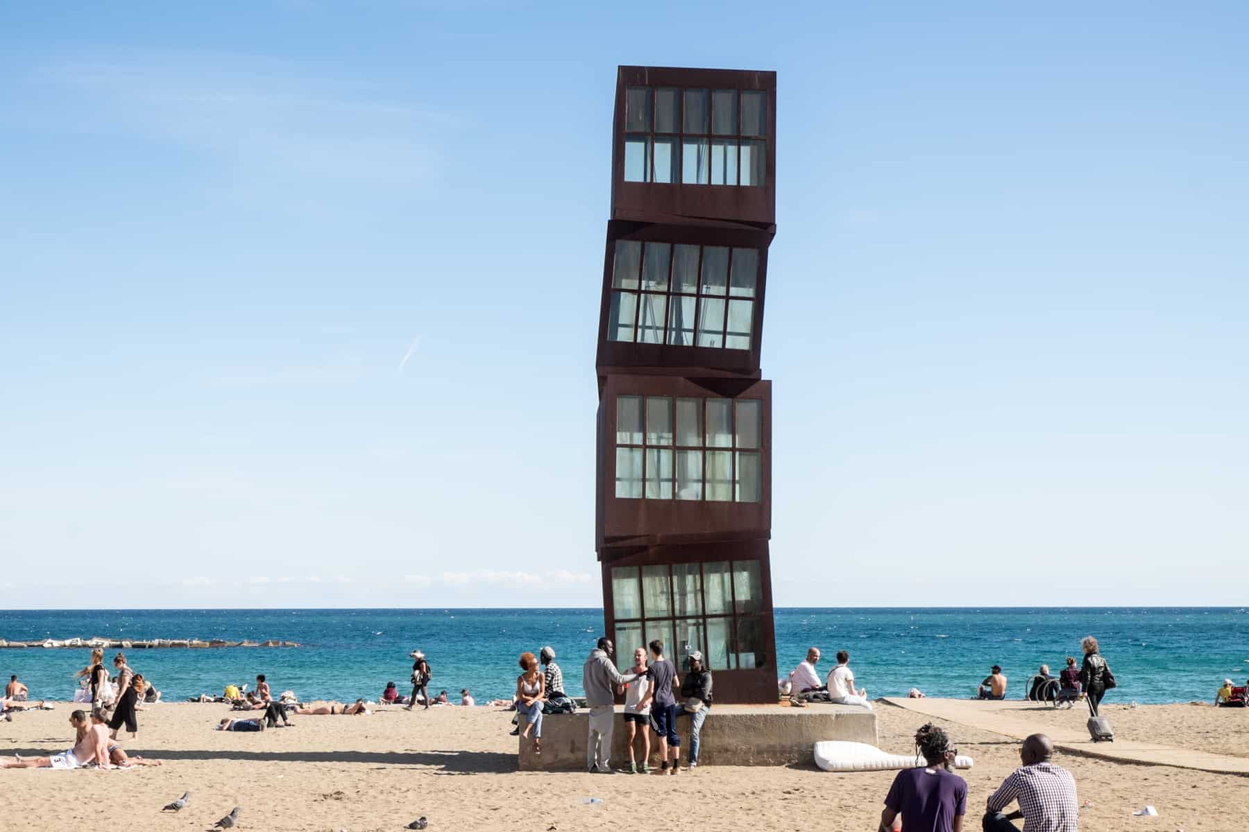 An artwork made of four, stacked brown squares with glass windows on the golden sand beach in Barcelona. The sea is a bright blue against a clear and sunny sky. 