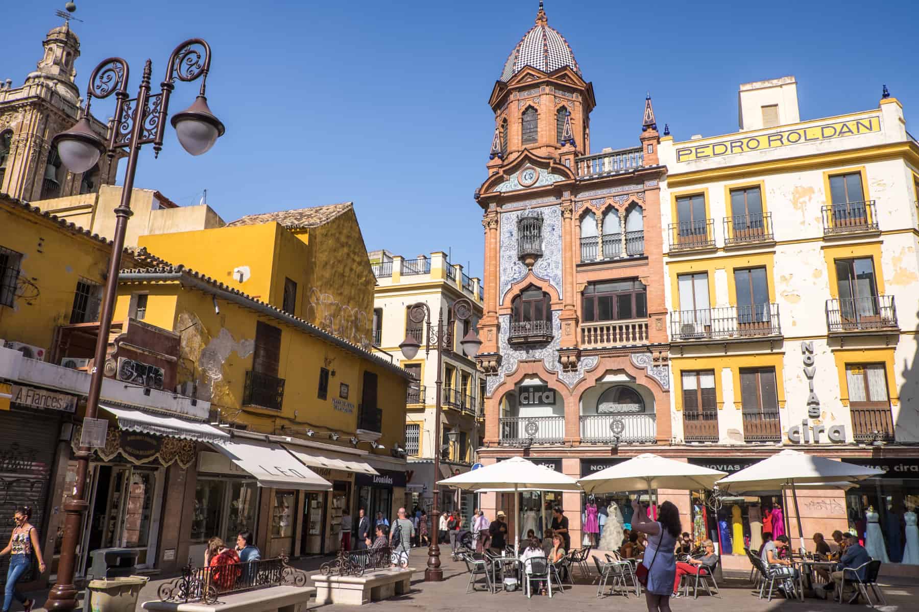 People walking through and sitting under white umbrellas in a public square in Seville Spain. The square is filled with buildings painted bright yellow and vivid salmon pink, vivid in the strong sunlight. 