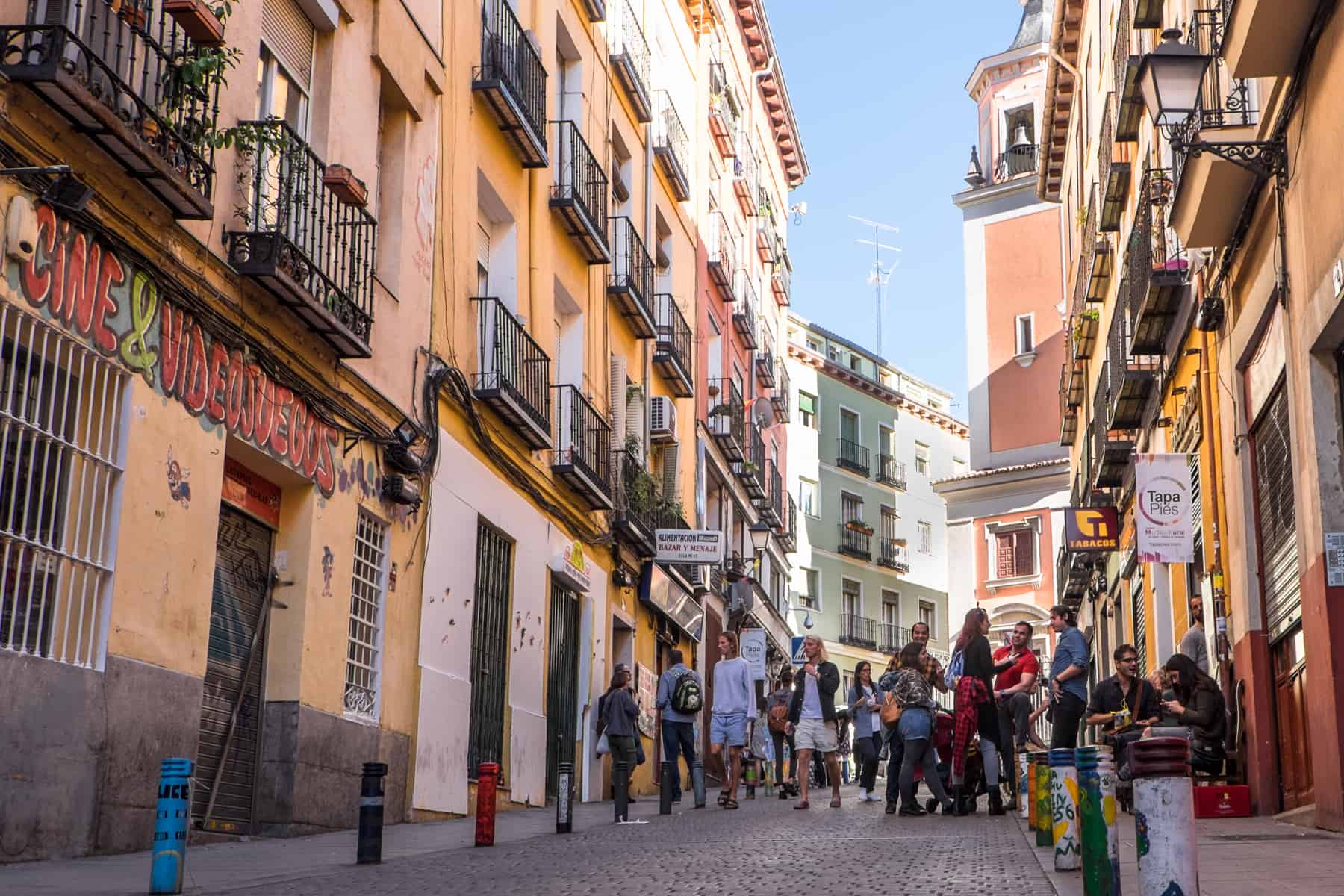 People sitting outside, standing and walking down a street in a neighbourhood in Madrid. The buildings are a mix of pastel colours and summery, butter yellows. 