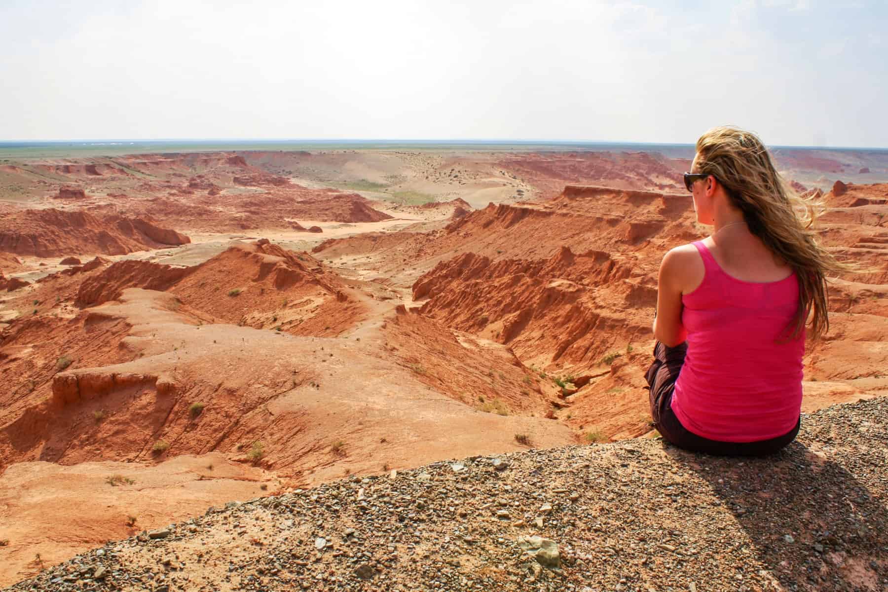 A woman sits if a rocky ledge overlooking the orange mars like landscape of Mongolia's Bayanzag Flaming Cliffs