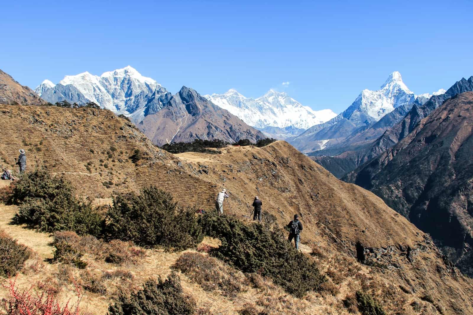 A small group of trekkers climbing to a higher altitude on golden mounds, on the Everest Base Camp trek. Three men can be seen in view taking photographs of the mountain range ahead.