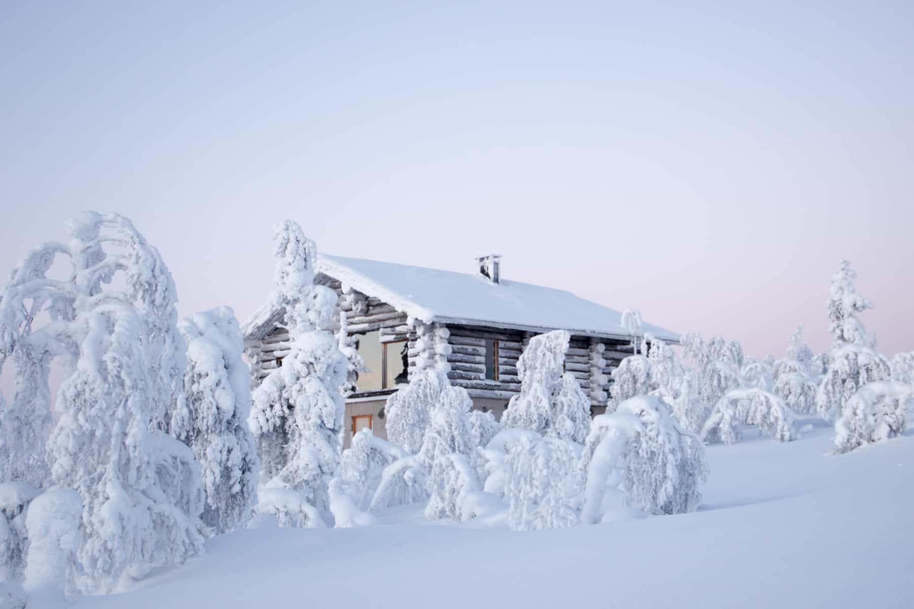A wooden house whose brown walls and roof are covered in snow, stands upon a thick mound of snow, surrounded by frozen trees. 