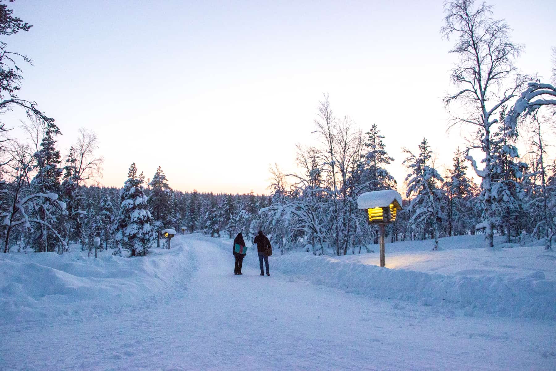 Two people walk down a wide pathway that cuts through a forest blanketed in snow, marked by a large square lamp with a yellow light. 