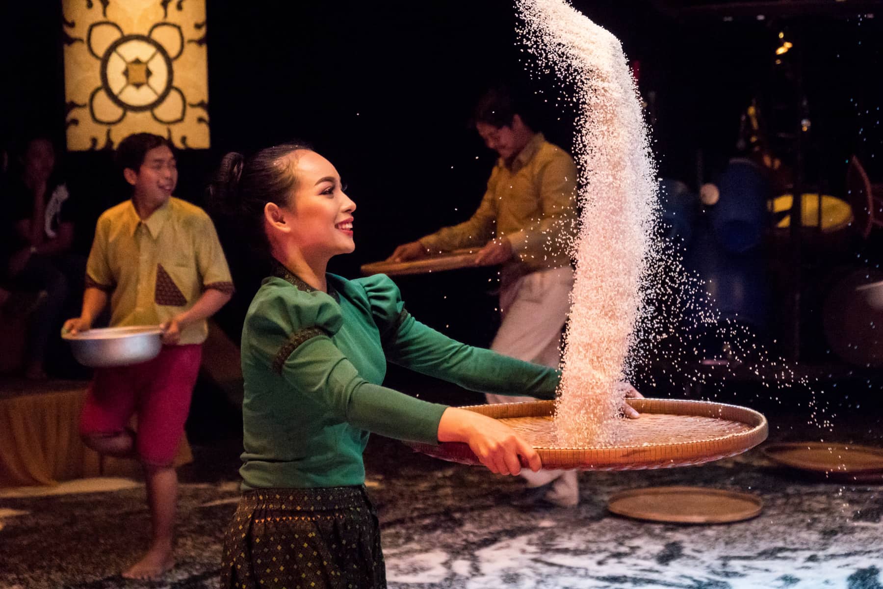 A Cambodian woman dressed in emerald green holds a wooden rice plate and decoratively throws a long line of rice into the air during a circus performance. Behind her are two men in yellow shirts holding bowls, and there is rice strewn all over the floor.
