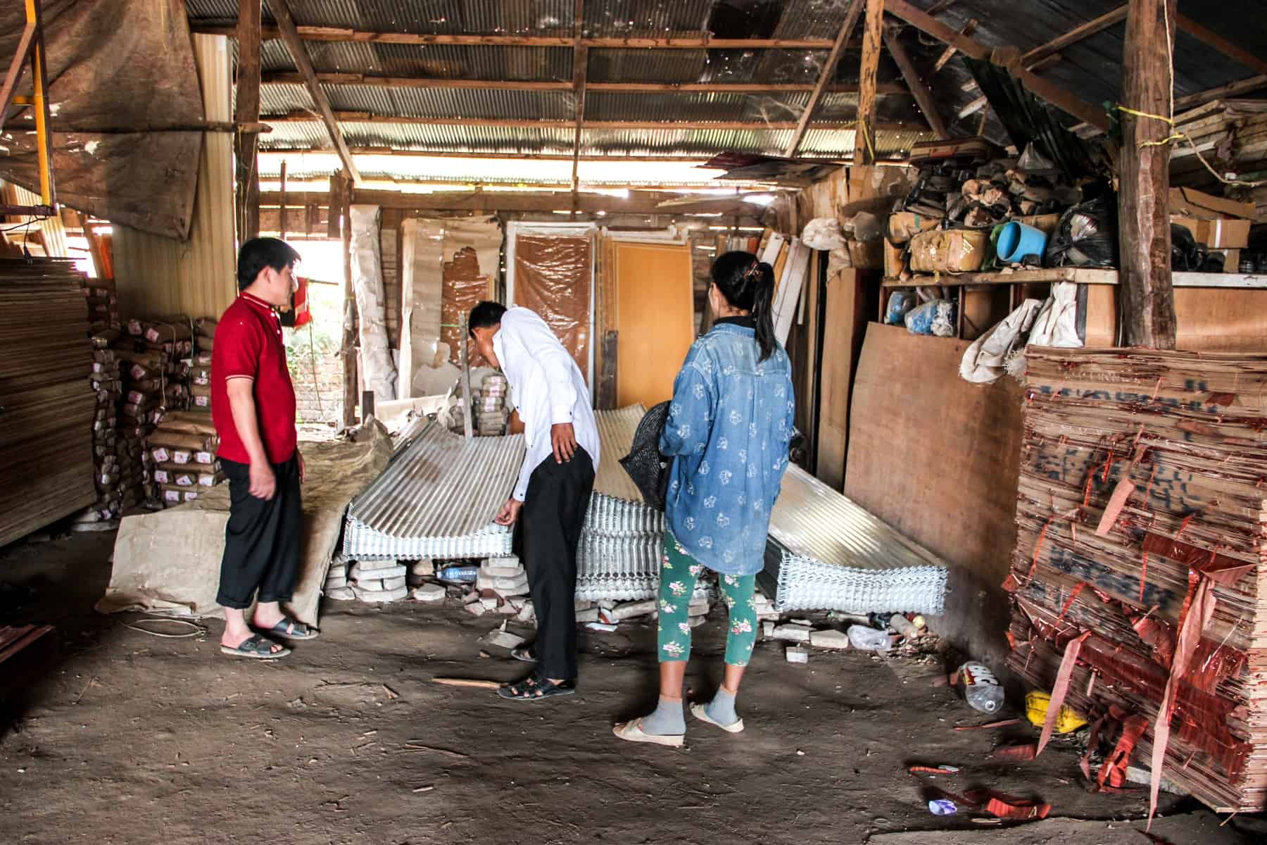 Three people inspect a pile of corrugated tin sheets inside a small wooden hut containing building materials.
