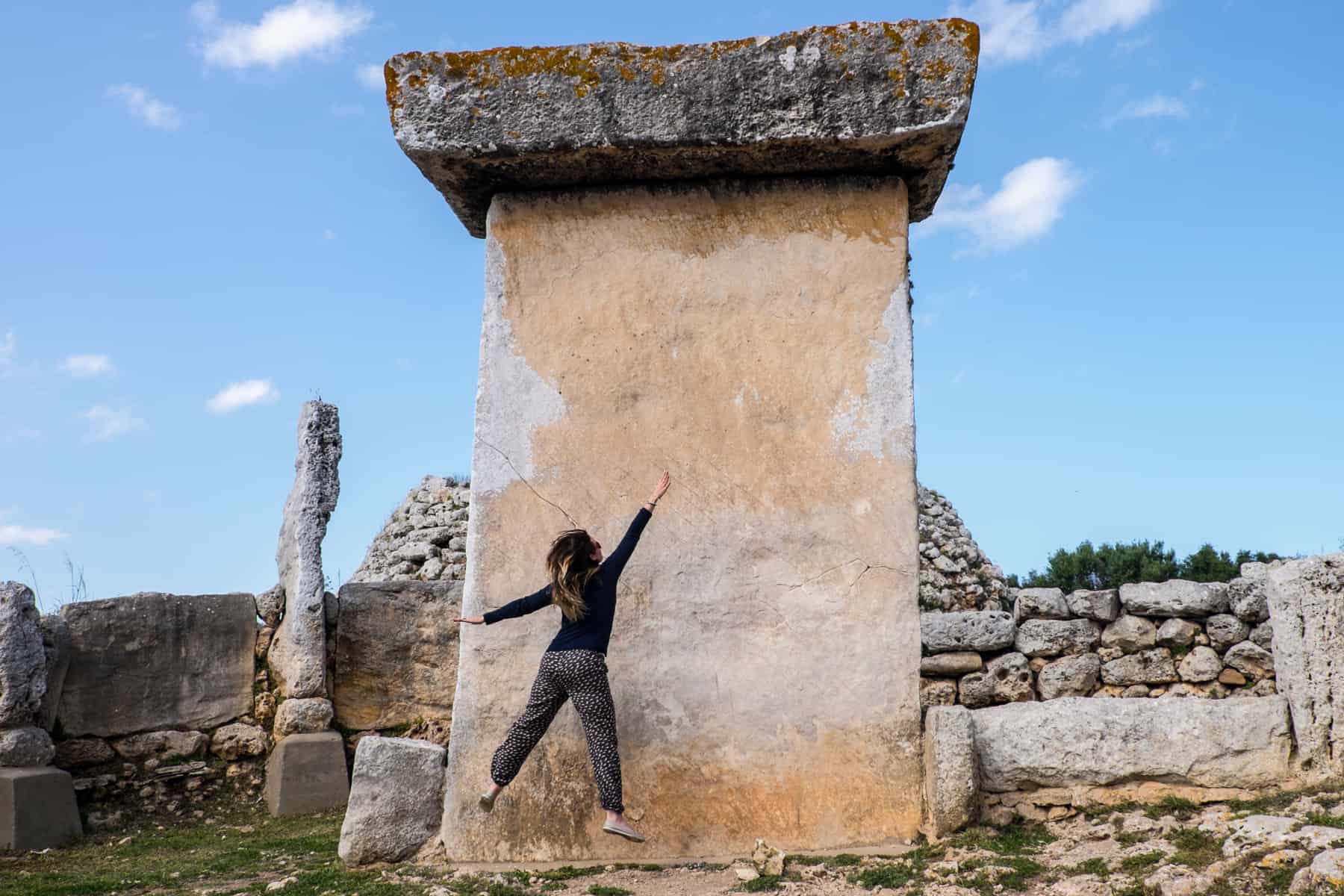 A woman jumping in front of a large golden rectangular rock with a flat rock roof - a prehistoric monument in Menorca, Balearic Islands. 
