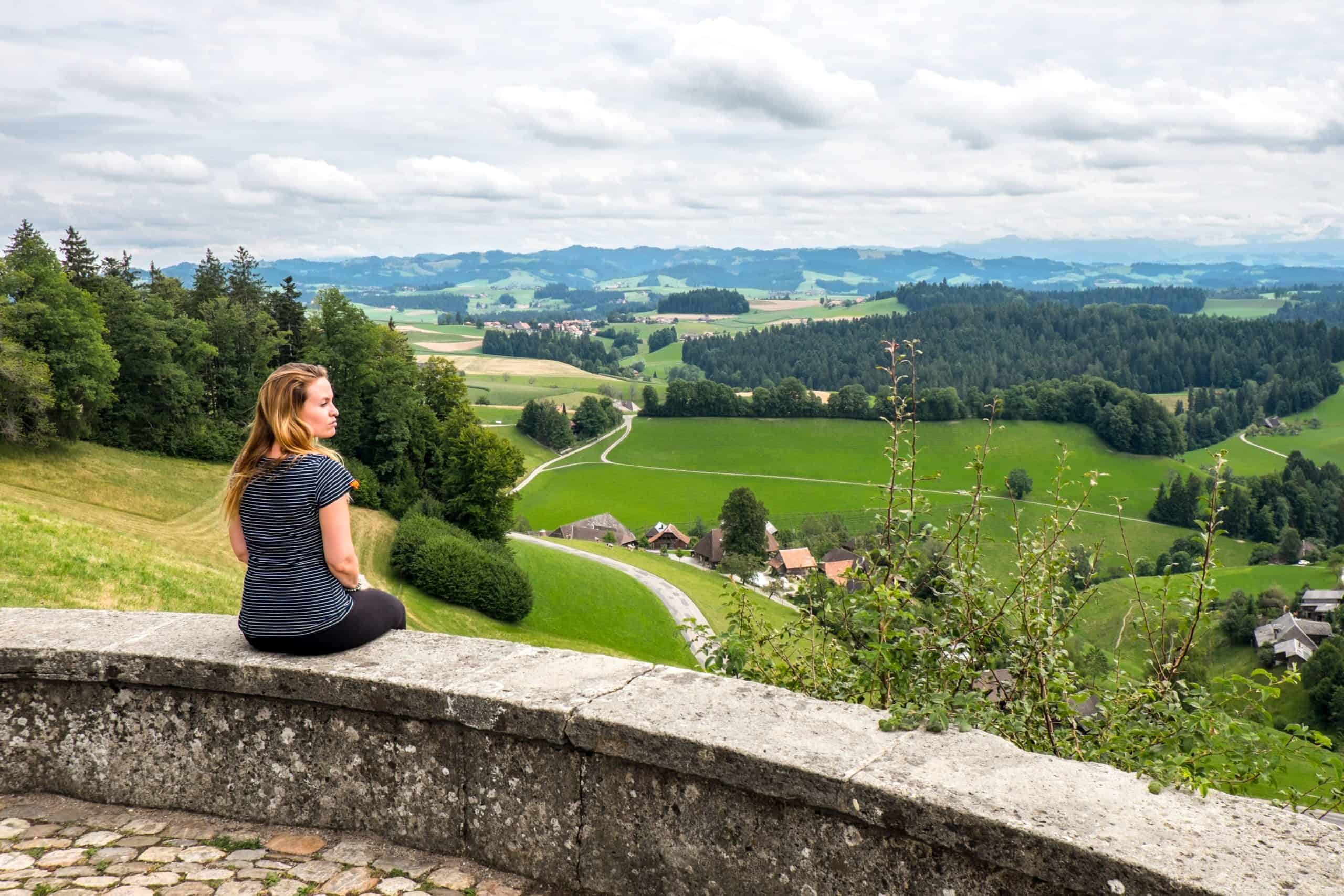 A woman sits on a stone wall across views of the Emmental Valley in Switzerland on the Cheese Route
