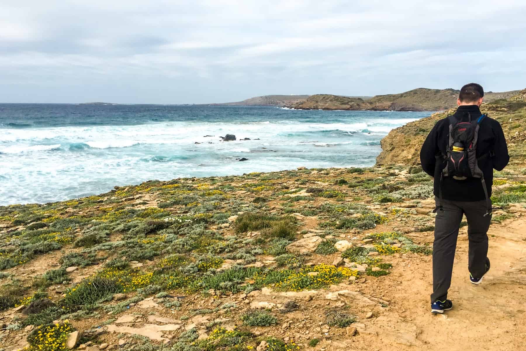 A man dressed in black walking the Cami de Cavalls in Menorca on a yellow, rocky trail next to the crashing waves of the sea. 
