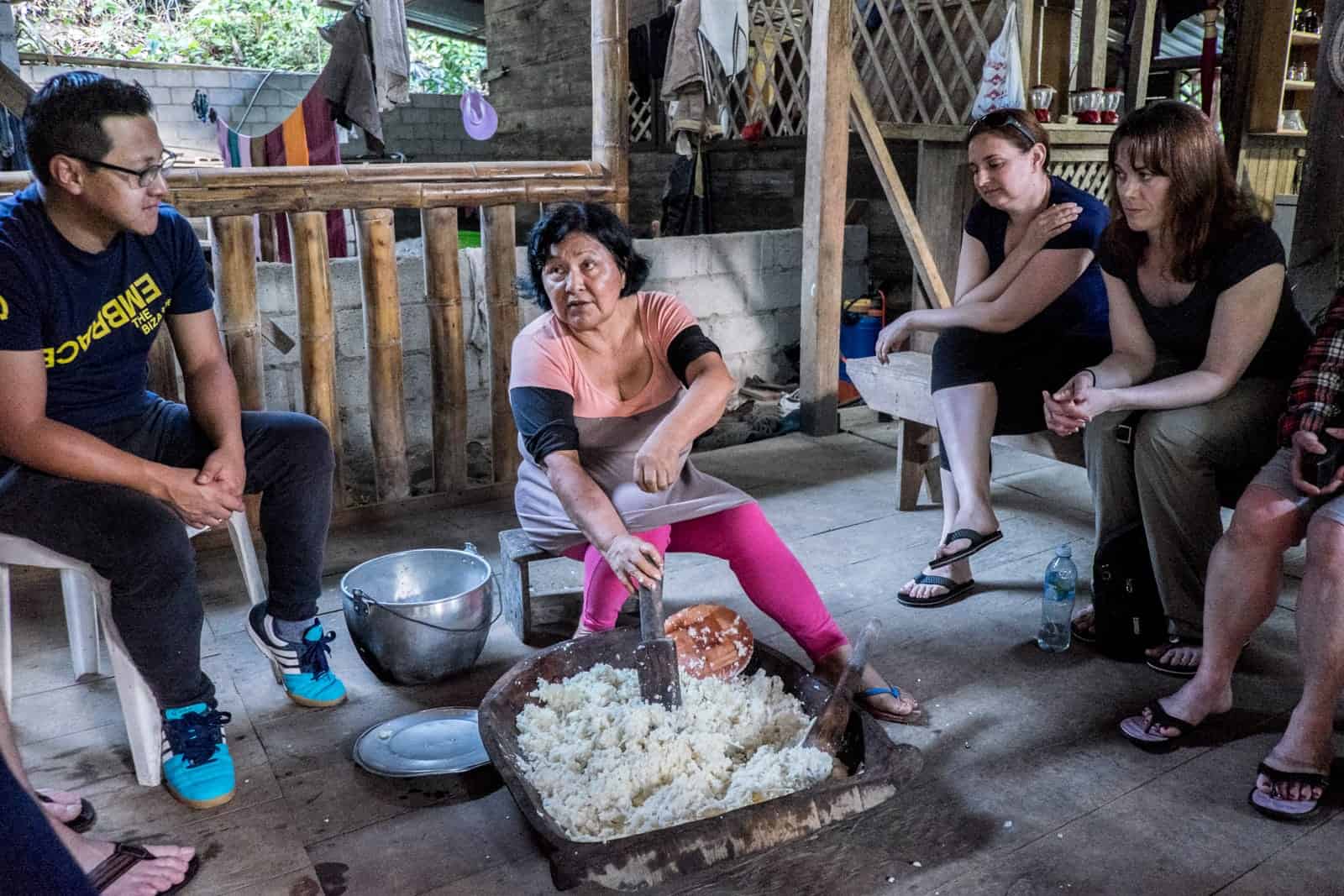 An Ecuadorian woman sitting on a wooden stall in a local house, stirs a thick, flour-like substance known as Chica, while travellers look on and learn. 