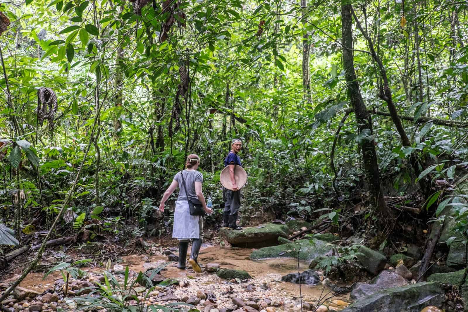 A woman follows a man carrying a large wooden bowel into the thick green foliage on a Jungle trek tour in the Ecuador Amazon.