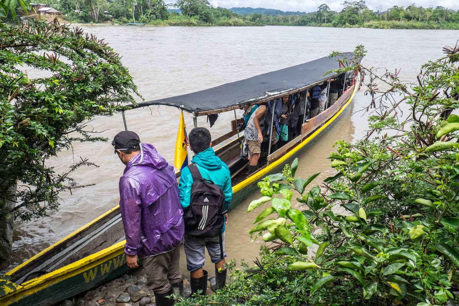 Passengers board a long, narrow yellow painted boat on the jungle shores of the Ecuador Amazon. The boat reaches out into the wide, muddy water. 