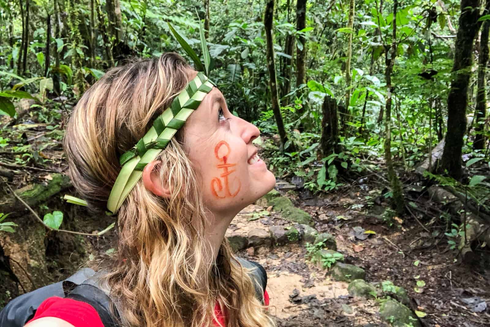 A woman with long blonde hair, wearing a headband made of woven plants and with a painted symbol on her face, stands within a jungle and looks up into the thick foliage.