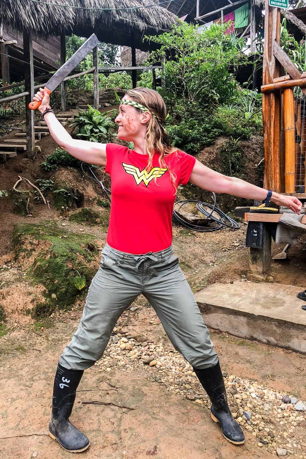 A woman in a red wonder woman t-shirt, khaki pants and black rubber boots stands in a fighting stance holding a machete in front of a wooden jungle lodge. 