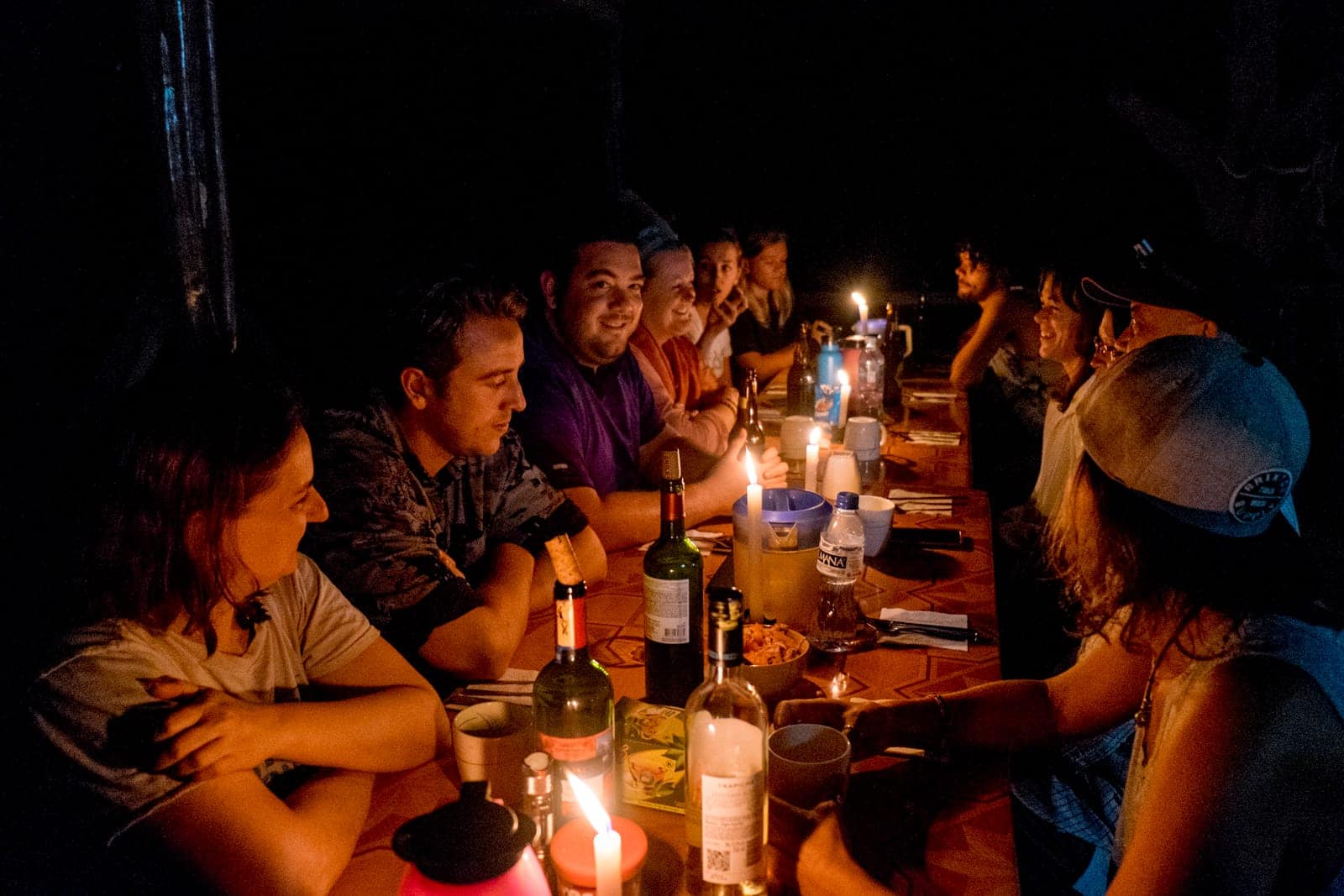 A group of people sitting around a table lined with bottles of wine and candle light, in the pitch black of the Amazon Rainforest.