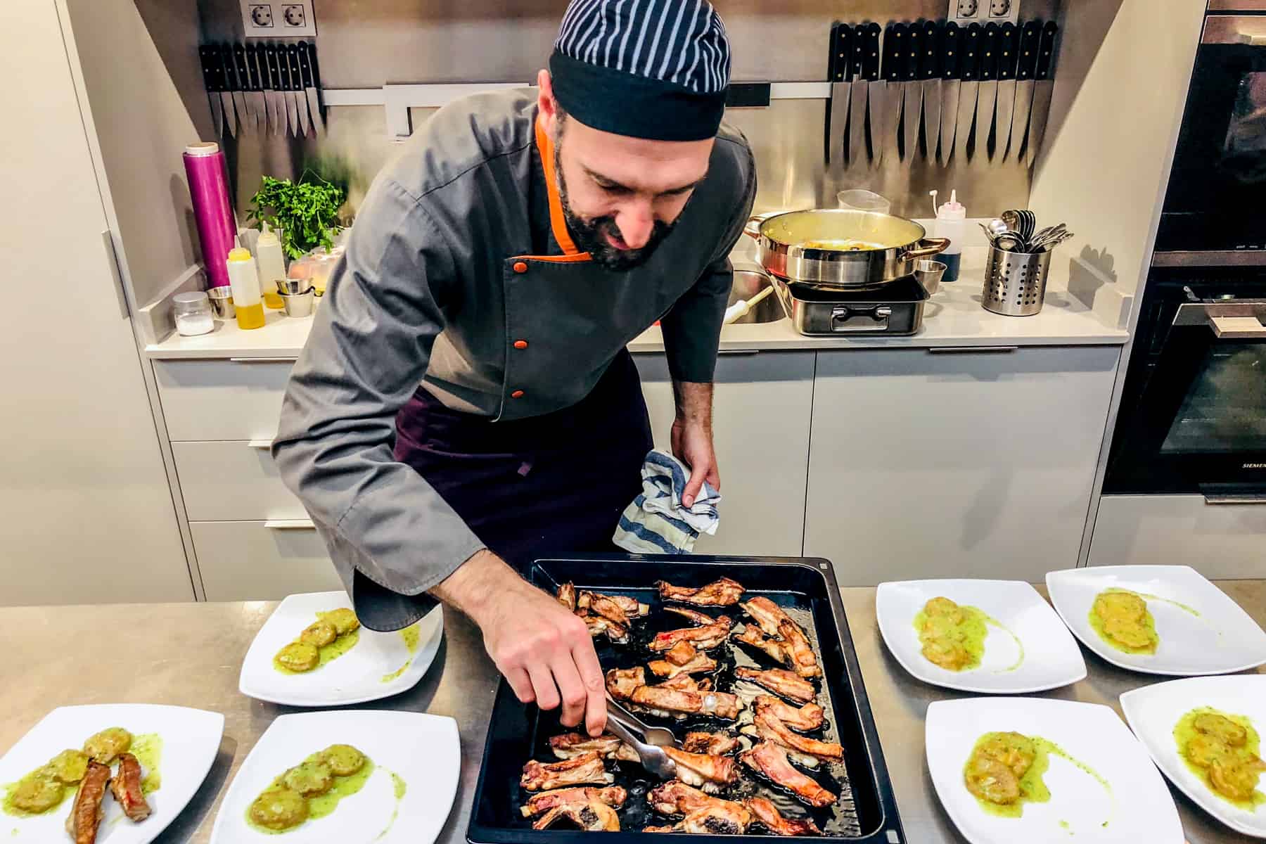 A man in a blue and white stripped hat and grey and orange chef's overalls, picks up grilled meat from a black tray, ready to place on the white plates featuring a green sauce. 