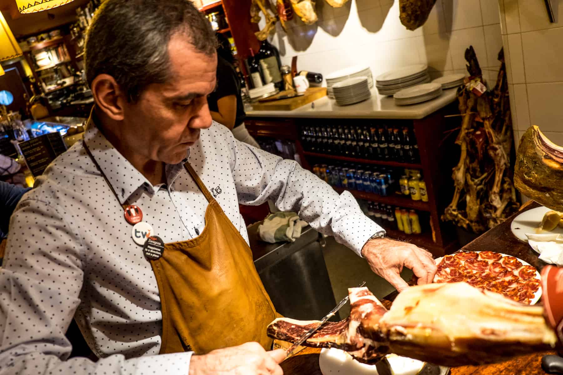 A man in a white doted shirt and brown apron, carves slices of red meat from a leg of Jamon. 