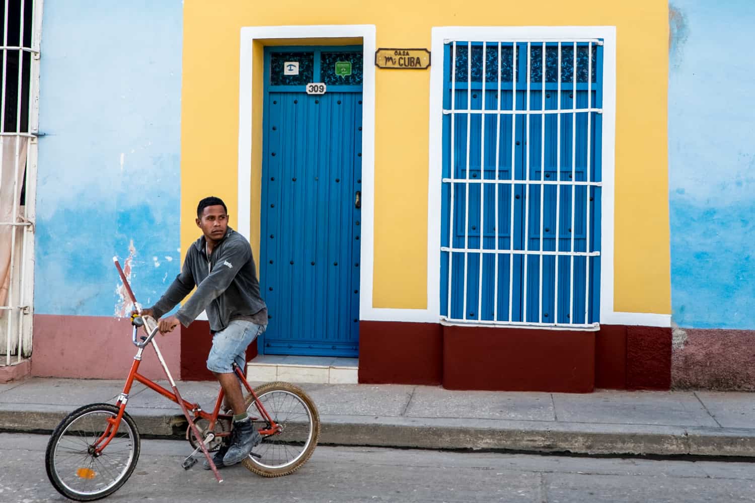 A man on a bike outside a blue and yellow painted Casa in Cuba. 