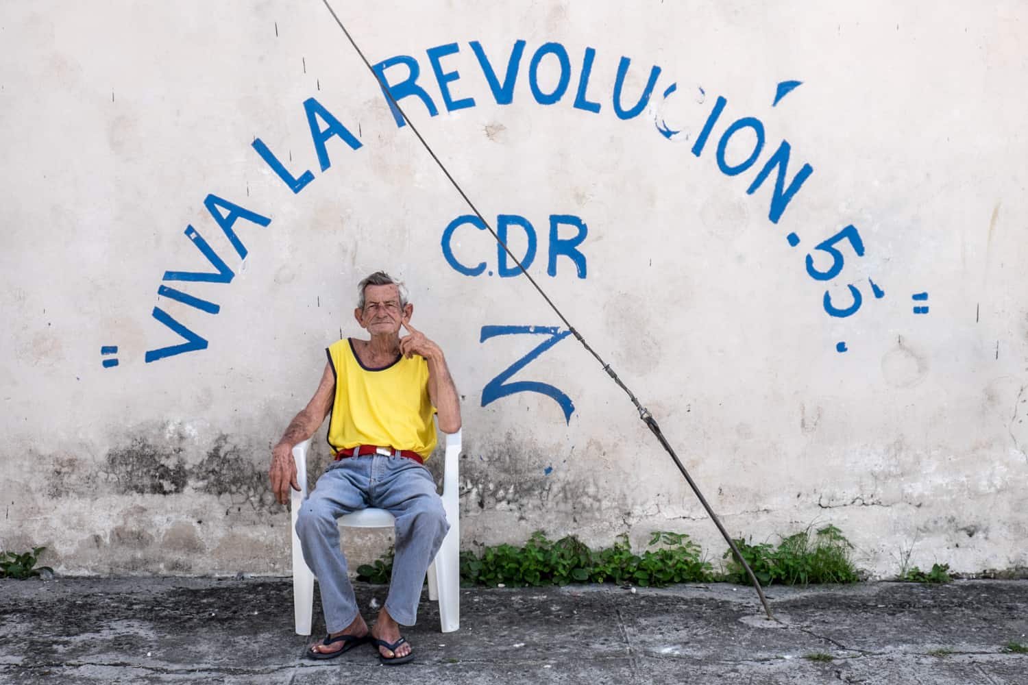 A Cuban man in a yellow vest sits in a white chair in front of a wall with the blue painted words: "Viva La Revolucion 55" CDR Z.