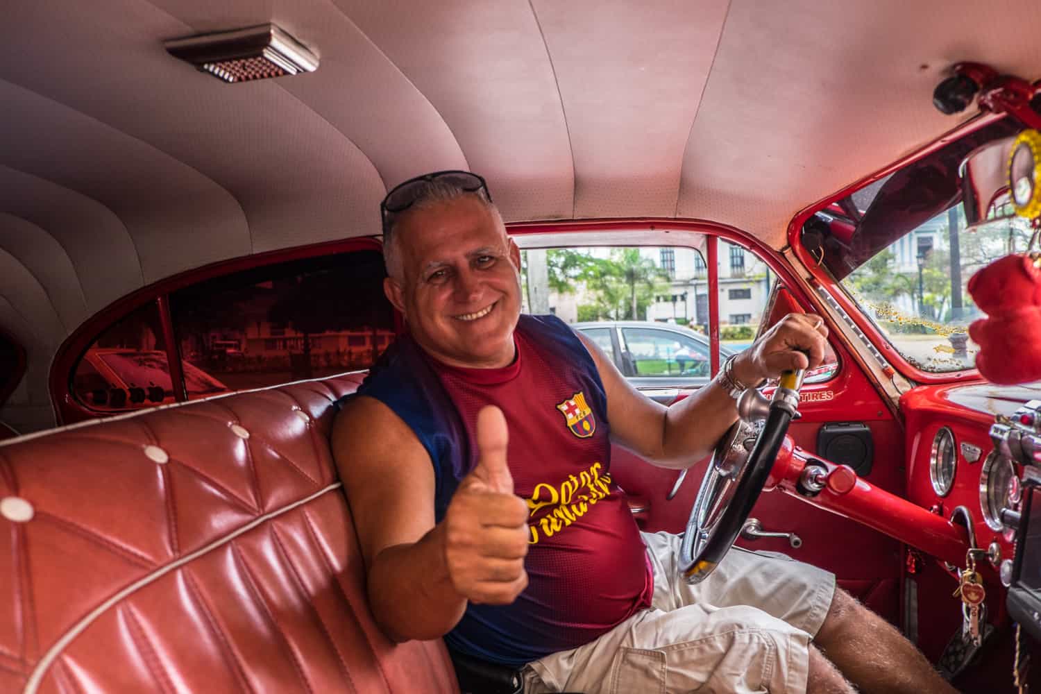 A man does a thumbs up sign from behind the wheel of a leather interior classic car in Cuba.