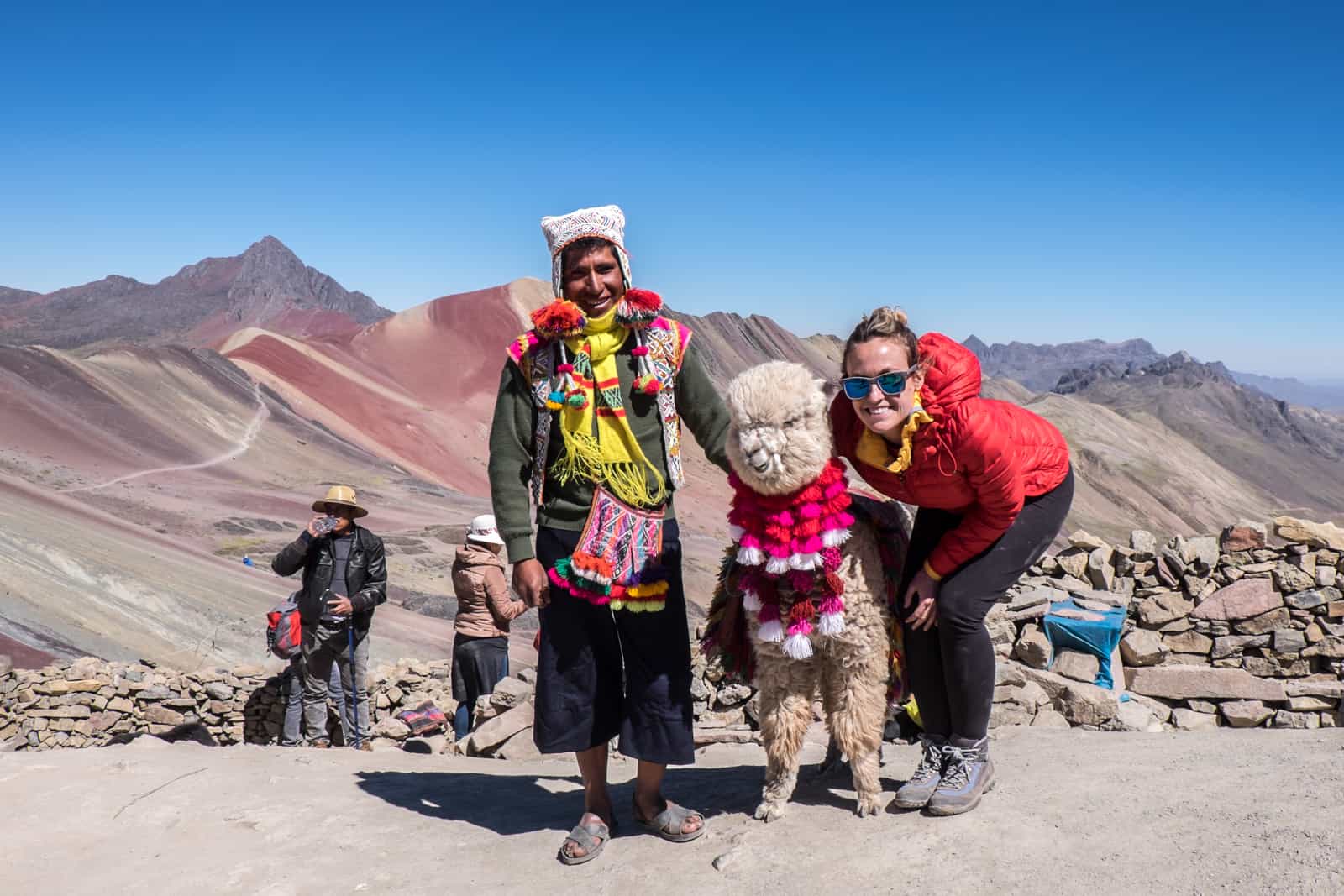 A tourist in Peru standing with a man and his alpaca at Rainbow Mountain (Vinicunca) in the Andes. 