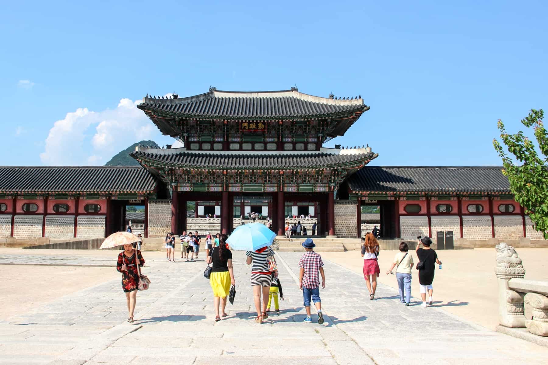 Tourists in Korea, some with sun umbrellas, walk towards the two-tier pagoda of Gyeongbokgung Palace in Seoul, South Korea. 