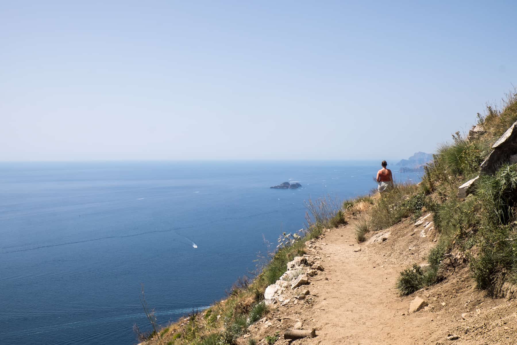 A woman walks ahead on a narrow yellow, forested cliff-edge trail overlooking the sea on the Path of the Gods. 