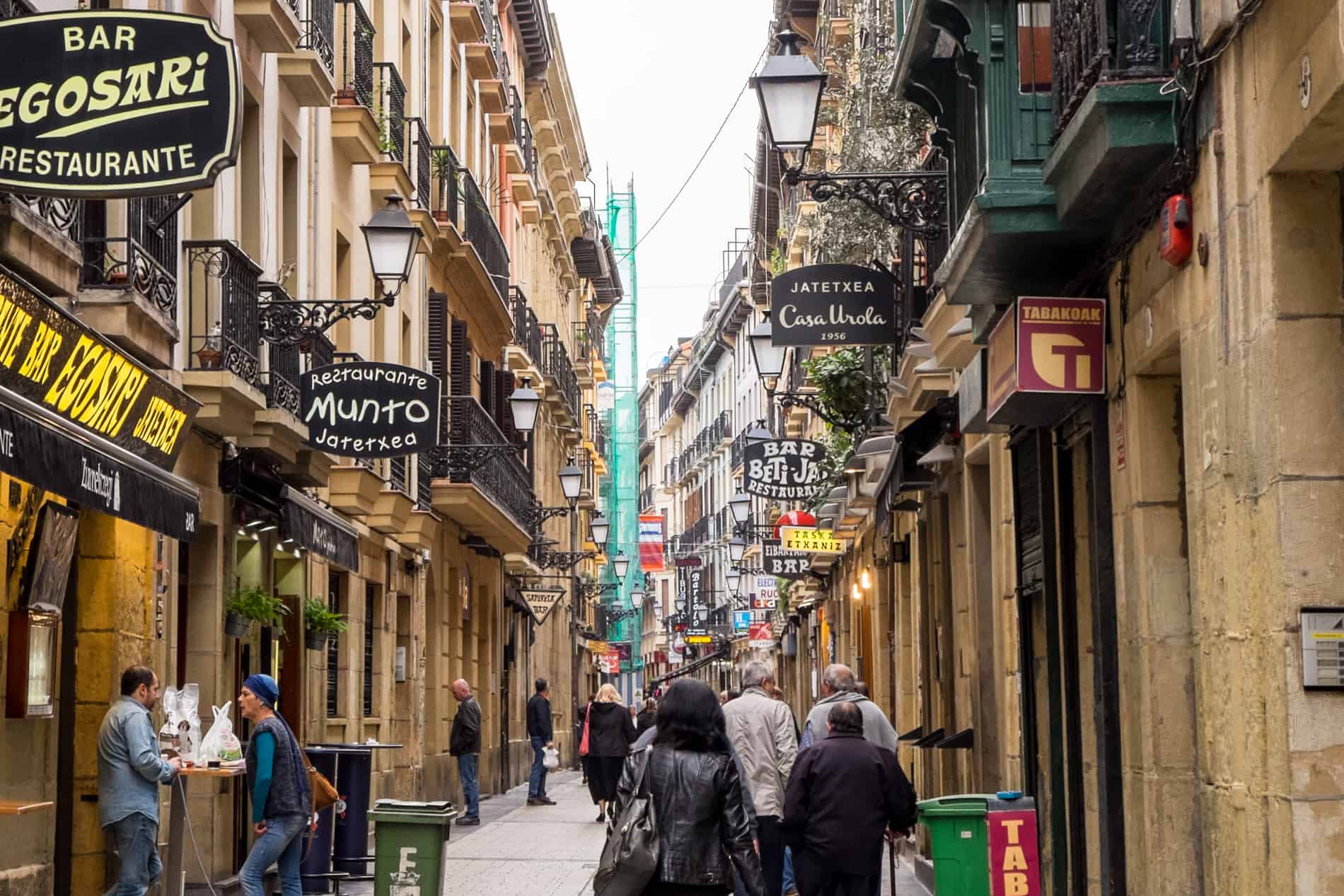 People walk down a golden walled street in San Sebastian old town lined with restaurant and Pintxos bar signs.