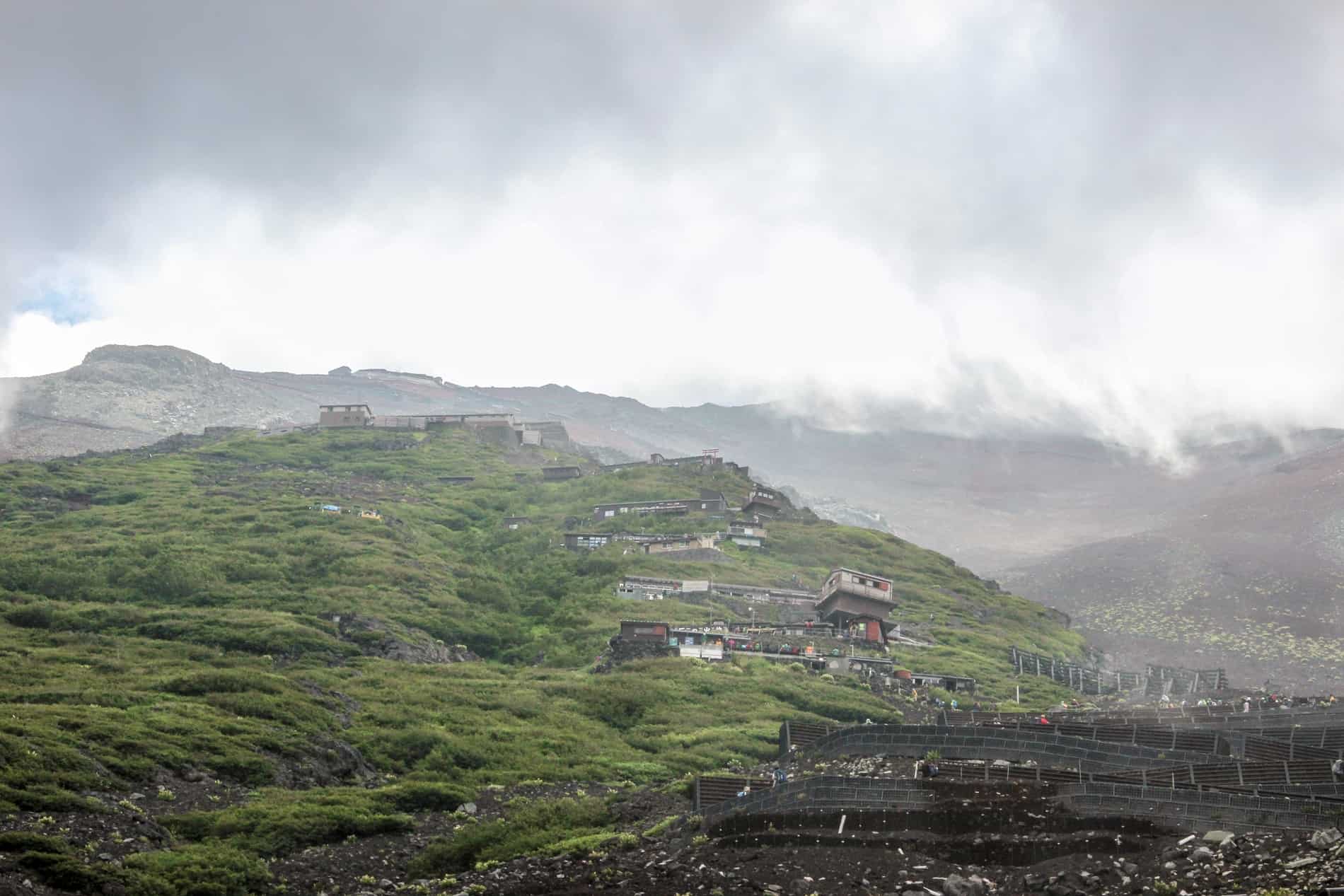 A Mt. Fuji hiking trail up a misty, rocky hill covered in green and a zigzag of mountain huts. 