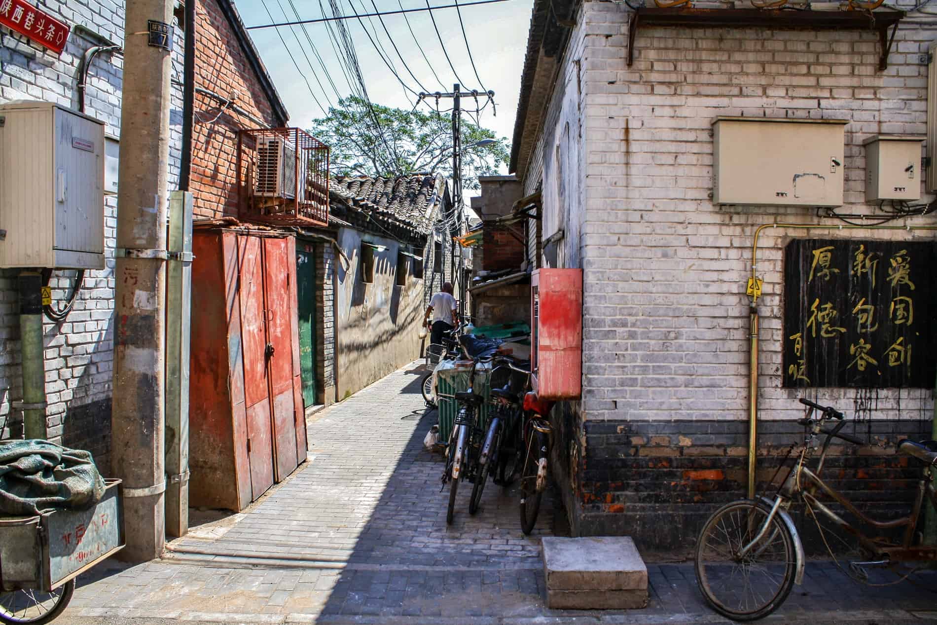A man walking down a street in a Beijing hutong lined with brick buildings and bikes. 